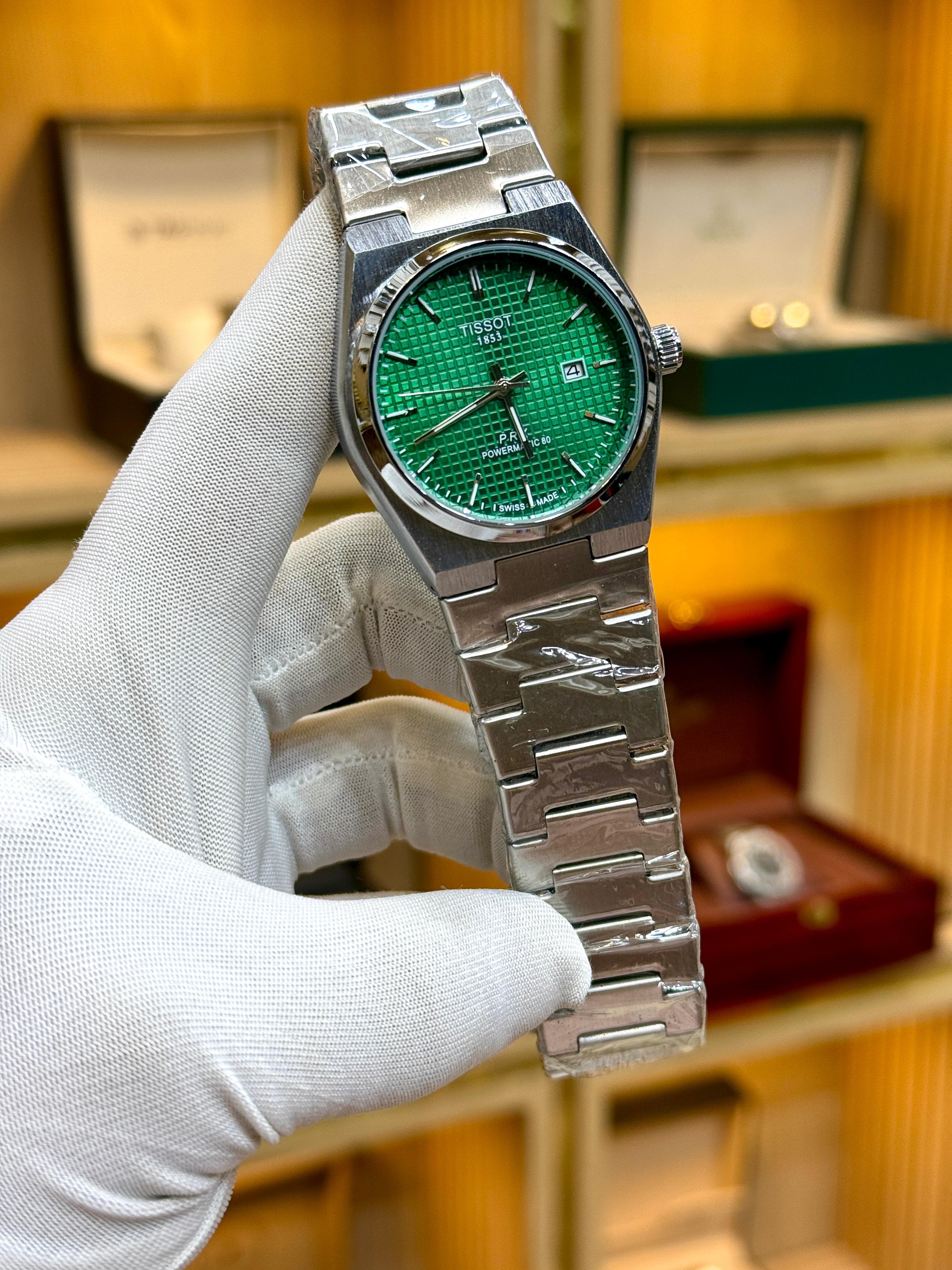 Silver watch with a green face held by a gloved hand against a blurred background