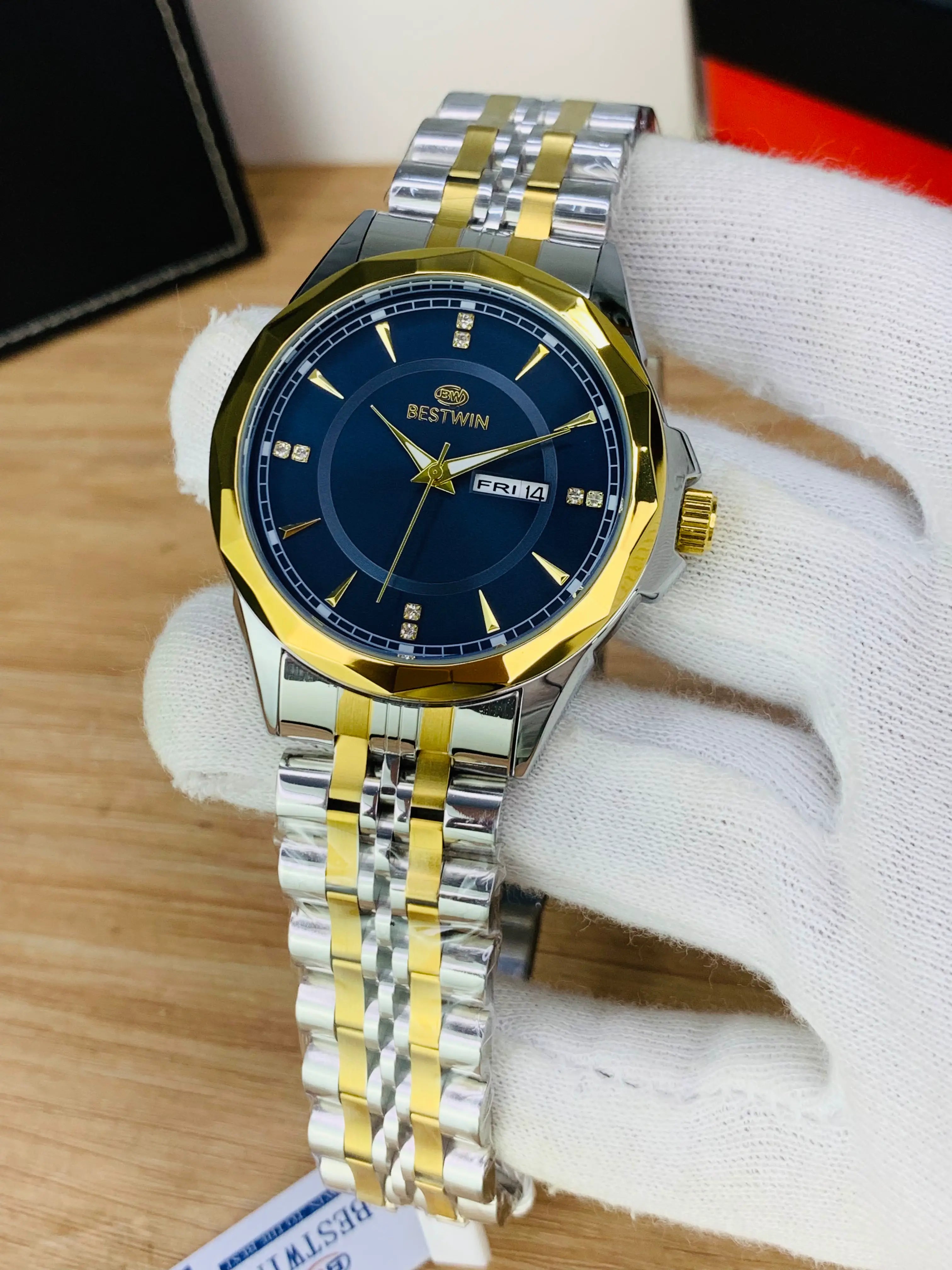 A two-tone stainless steel and gold Bestwin wristwatch with a deep blue dial, held in a white gloved hand over a light wood surface.
