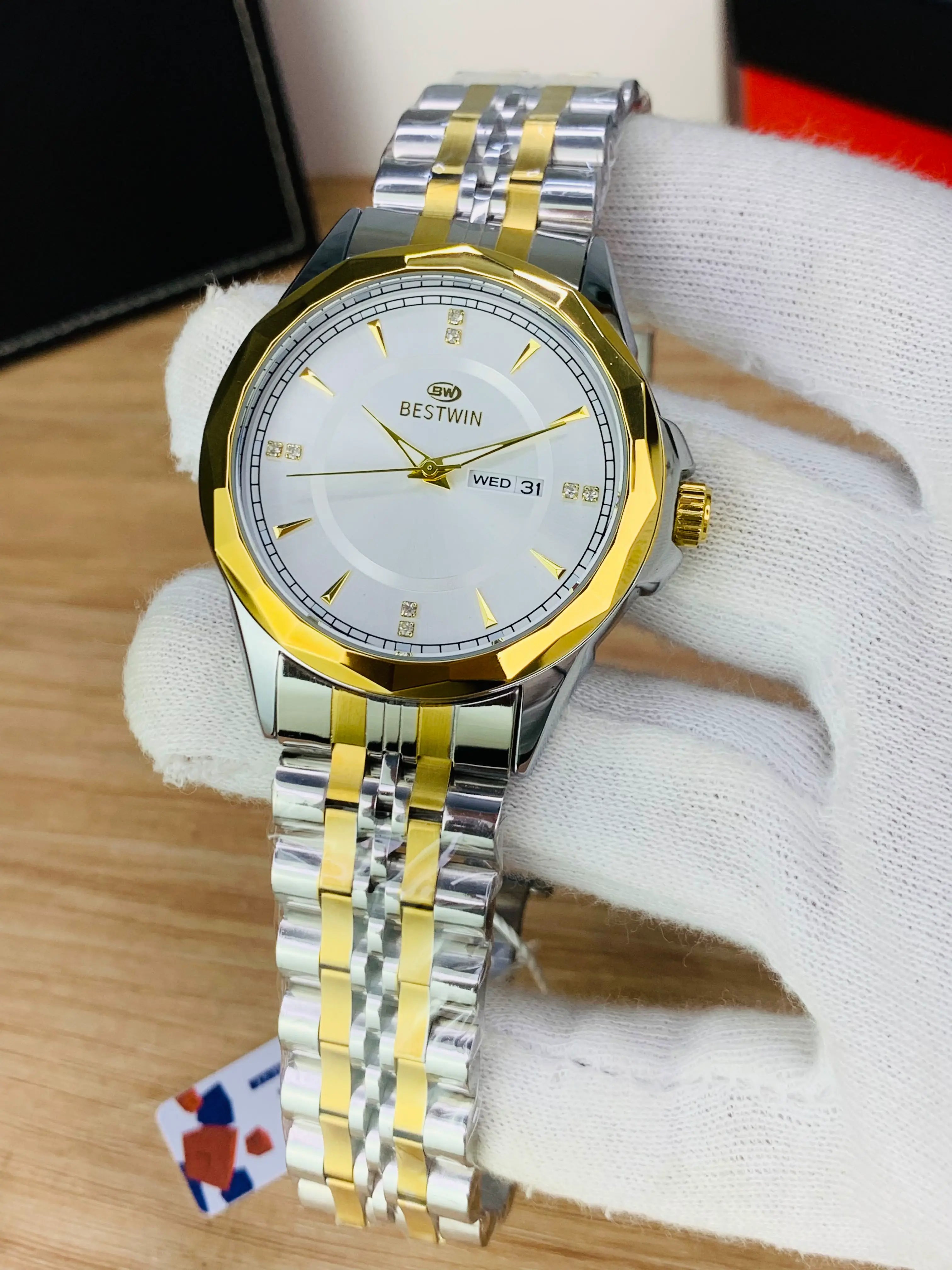 A two-tone stainless steel and gold Bestwin wristwatch with a white dial, held in a white-gloved hand over a light wood surface.