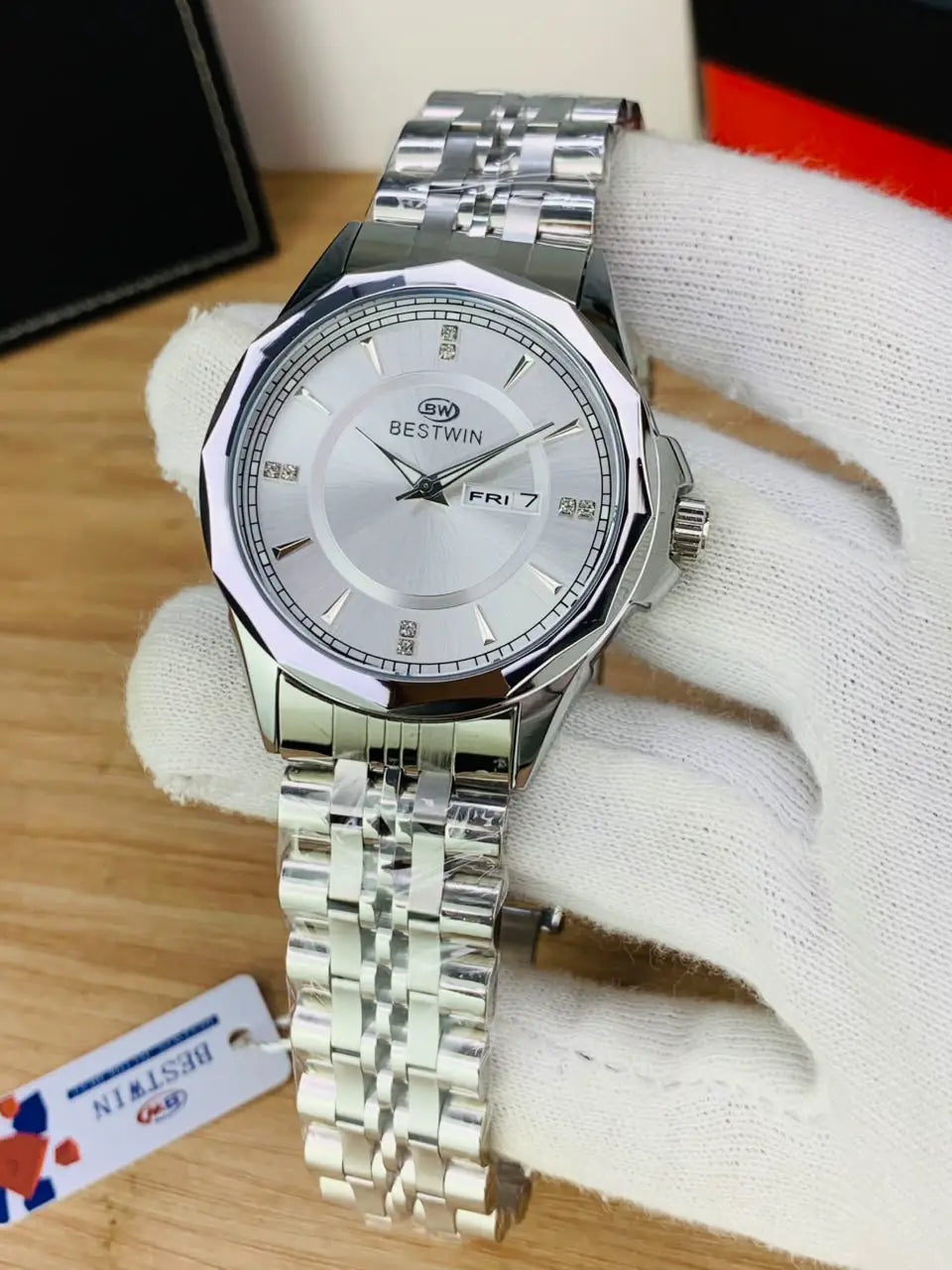 A silver Bestwin wristwatch with a stainless steel link bracelet, held in a white-gloved hand over a wooden surface. 